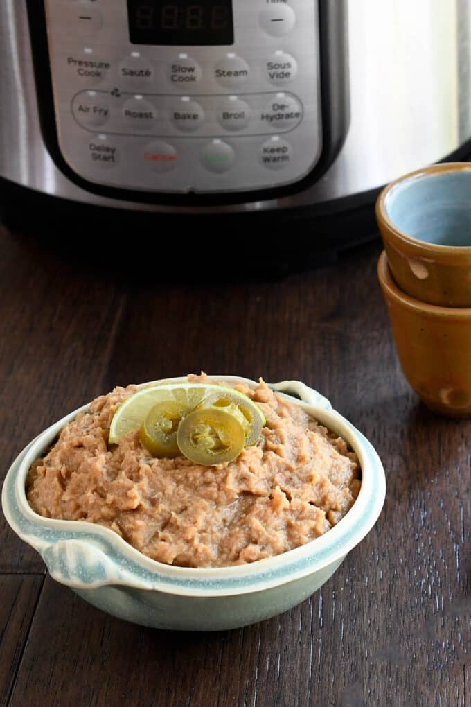 Light blue bowl of refried beans topped with pickled jalapenos and lime slice, in front of an instant pot.