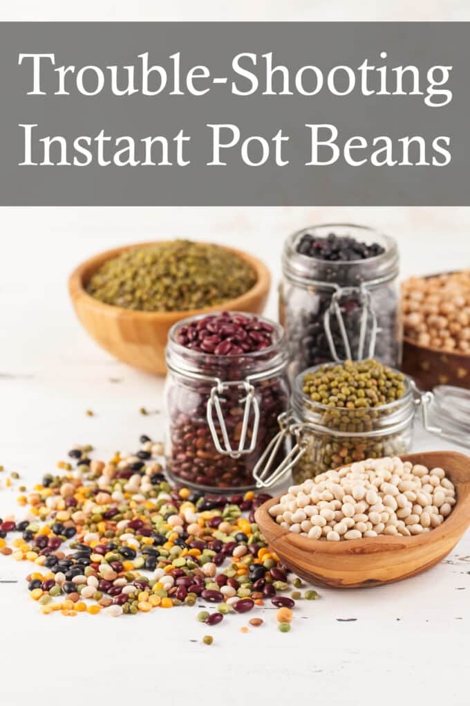 Several jars of different beans, and several wooden bowls of different beans on a white table with a variety of beans scattered on the table.