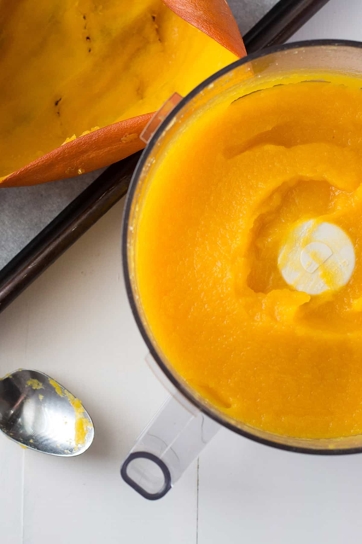 A food processor with pureed pumkin in it. Behind it is the hallowed out skin of the pumpkin.