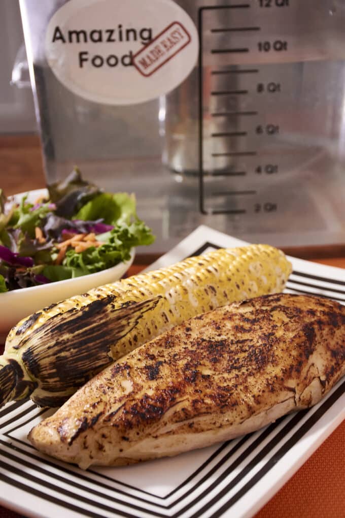 Grilled chicken breast and corn on the cob on a white and black plate. Salad and sous vide machine in background.