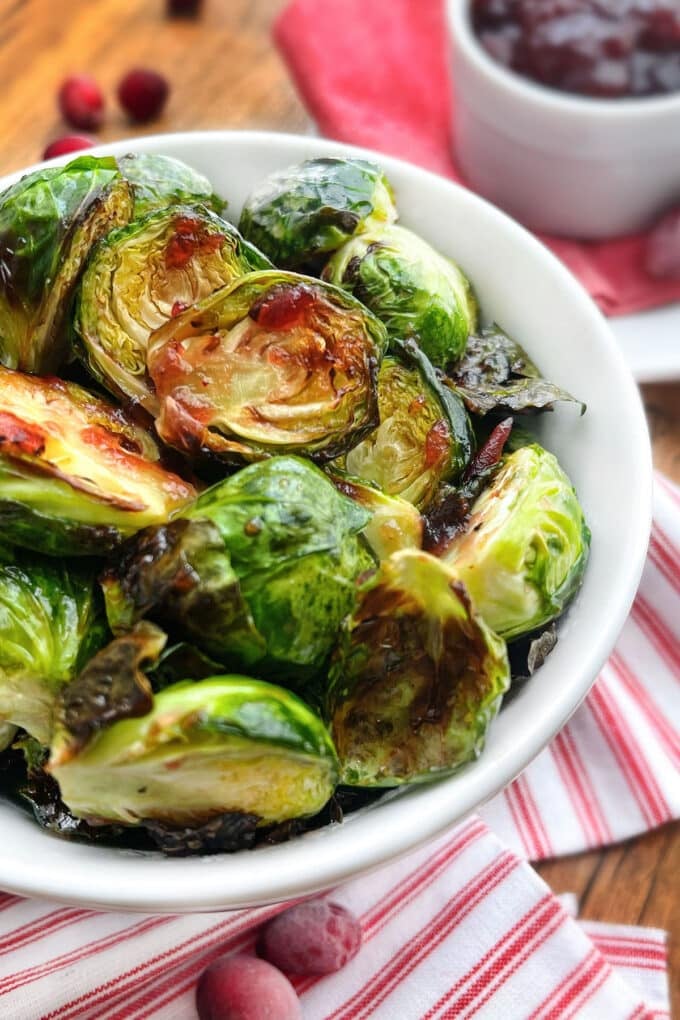 Brussels Sprouts with cranberry glaze in a white bowl, cranberries and sauce in background.