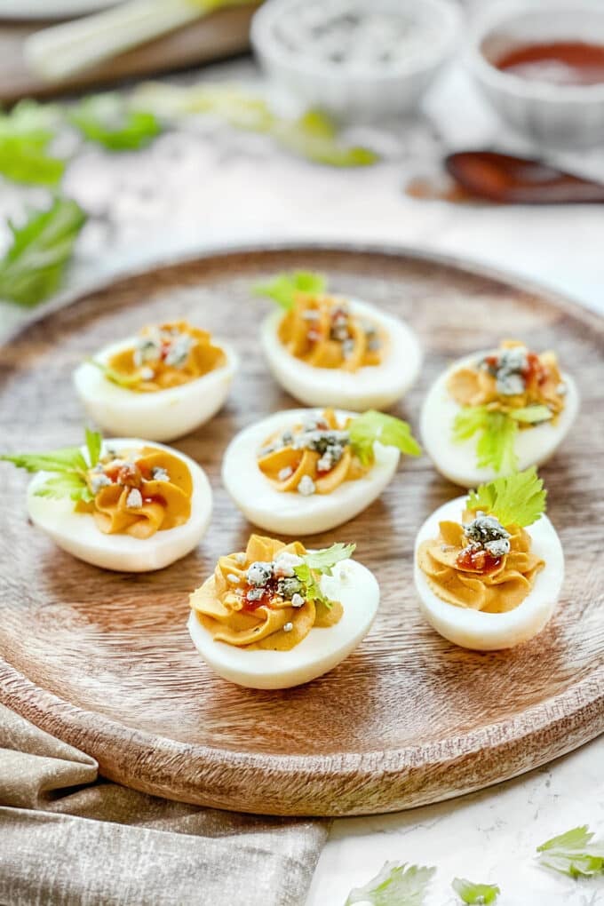 Buffalo Deviled Eggs topped with bleu cheese and buffalo sauce on a round wooden platter.