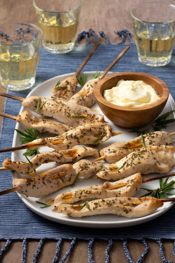 Chicken Skewers with Rosemary, on a white plate with a brown bowl of aioli, glasses of white wine in background.