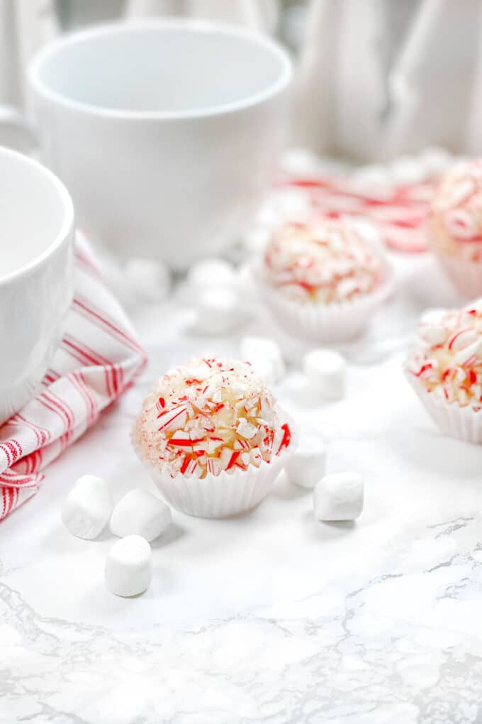 White Chocolate Cocoa Bombs covered in crushed candy cane, next to white mugs and marshmallows.