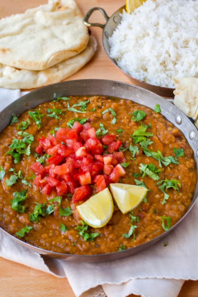 Red lentil dal in a pan, topped with diced tomatoes and parsley. Naan and white rice in background.