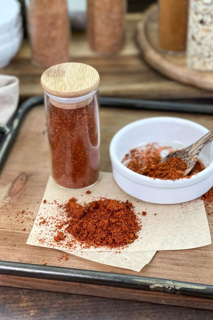 Deep red Tajin seasoning blend in a glass spice jar, white dish, and spilled on parchment.