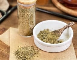 Homemade poultry seasoning in glass jar, in dish with spoon, and spilled on parchment paper.