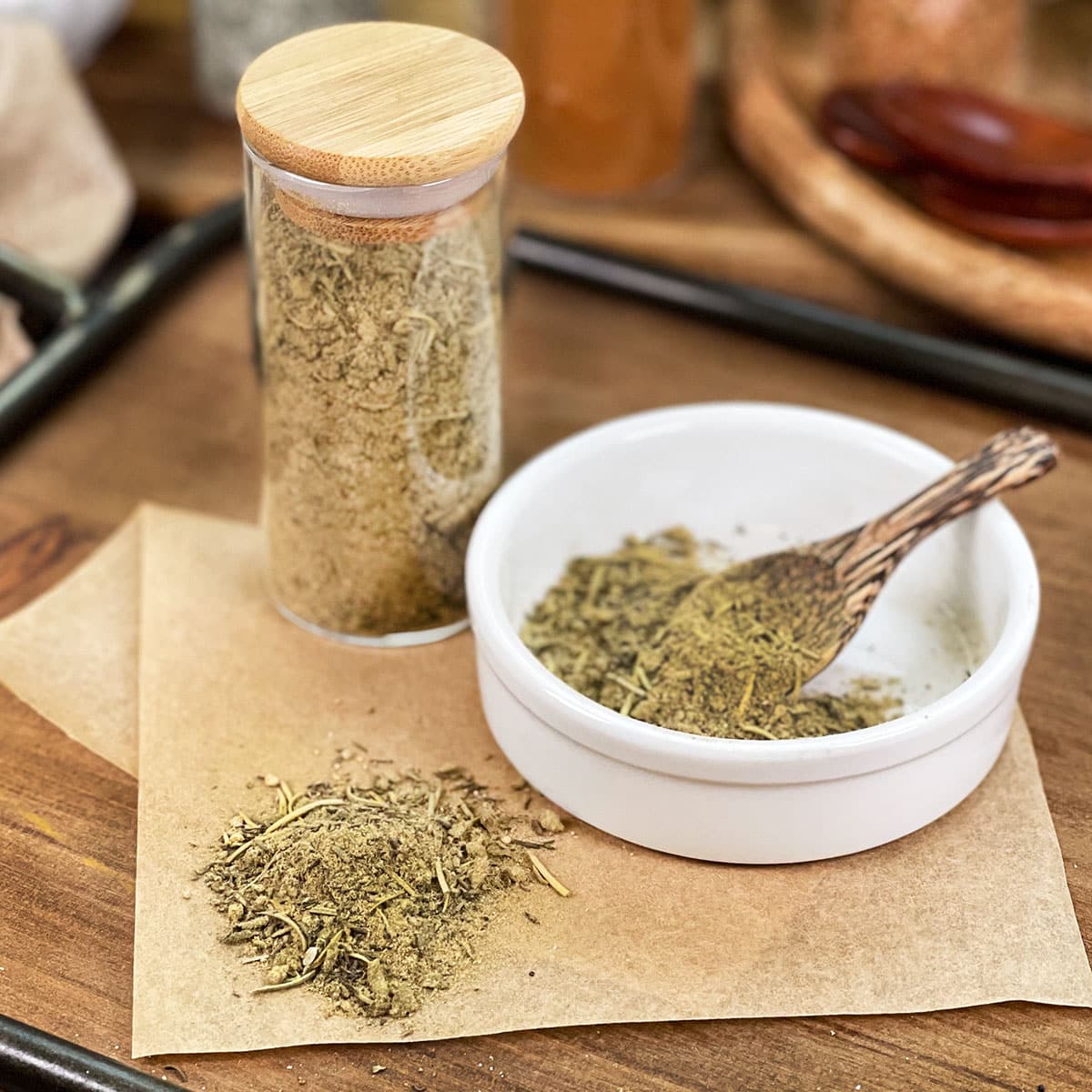 Homemade poultry seasoning in glass jar, in dish with spoon, and spilled on parchment paper.