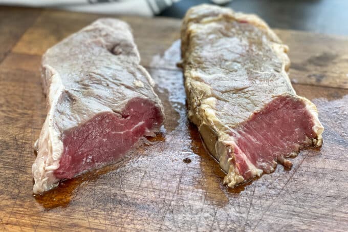 Two raw and marinated NY strip steaks on a wooden cutting board.