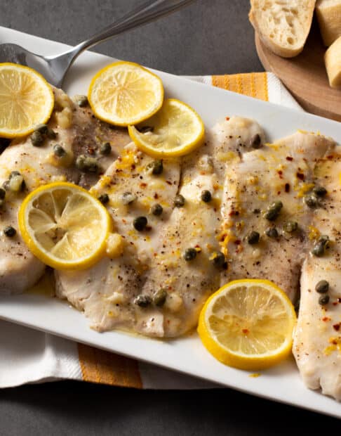 Baked tilapia fillets with capers and lemon slices on a white platter.