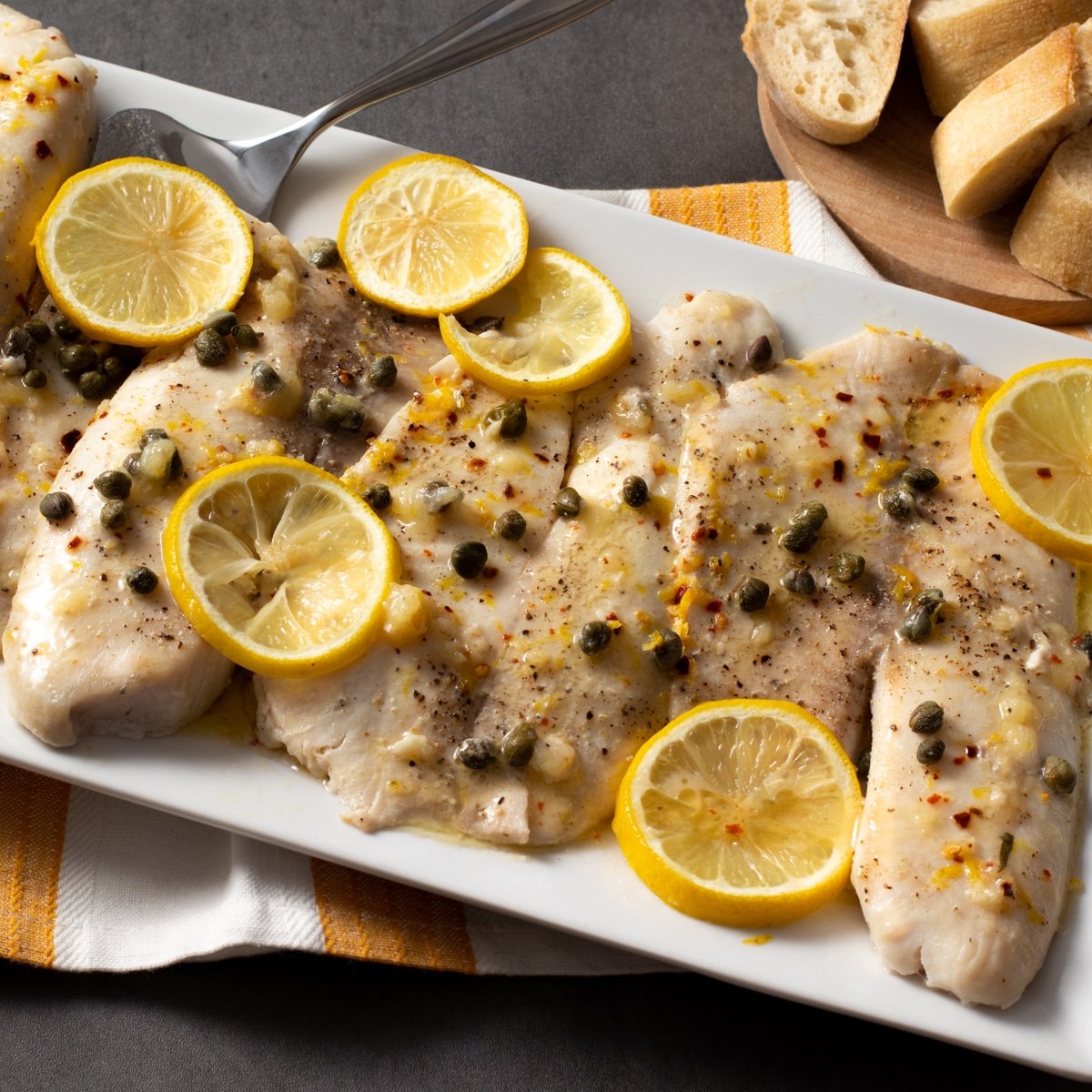 Baked tilapia fillets with capers and lemon slices on a white platter.