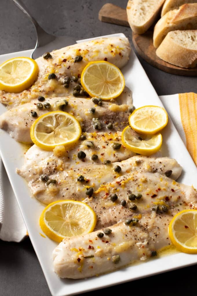 Baked tilapia fillets with capers and lemon slices on a white platter.