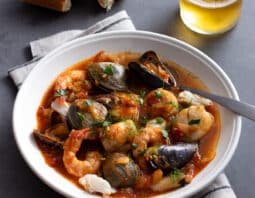 Seafood stew with fish, clams, shrimp, and more in a tomato based broth in a white bowl.