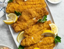 Fried catfish fillets with cornmeal coating on a white platter with lemon wedges.