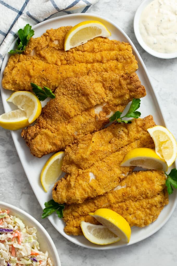 Fried catfish fillets with cornmeal coating on a white platter with lemon wedges.