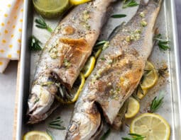 Two whole branzino on a sheet pan with sliced lemon and herbs.