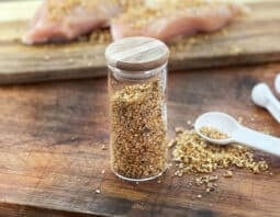 Small glass jar of montreal seasoning blend, raw seasoned chicken in background.