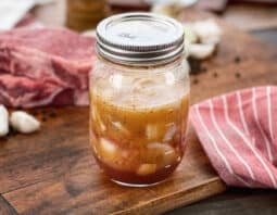 Mason jar with steak marinade with onion and garlic, raw steak in background.