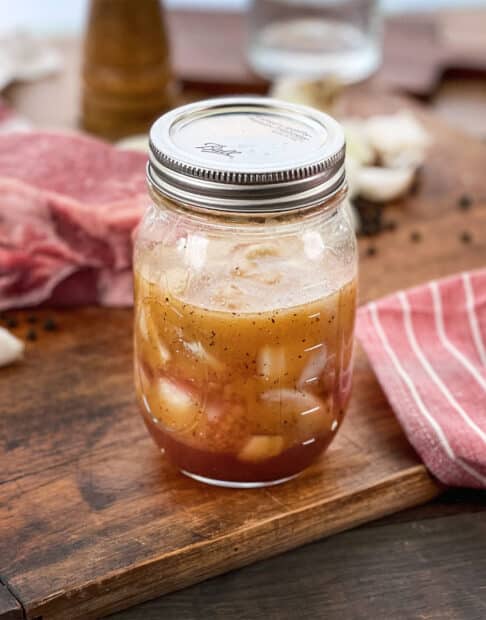 Mason jar with steak marinade with onion and garlic, raw steak in background.