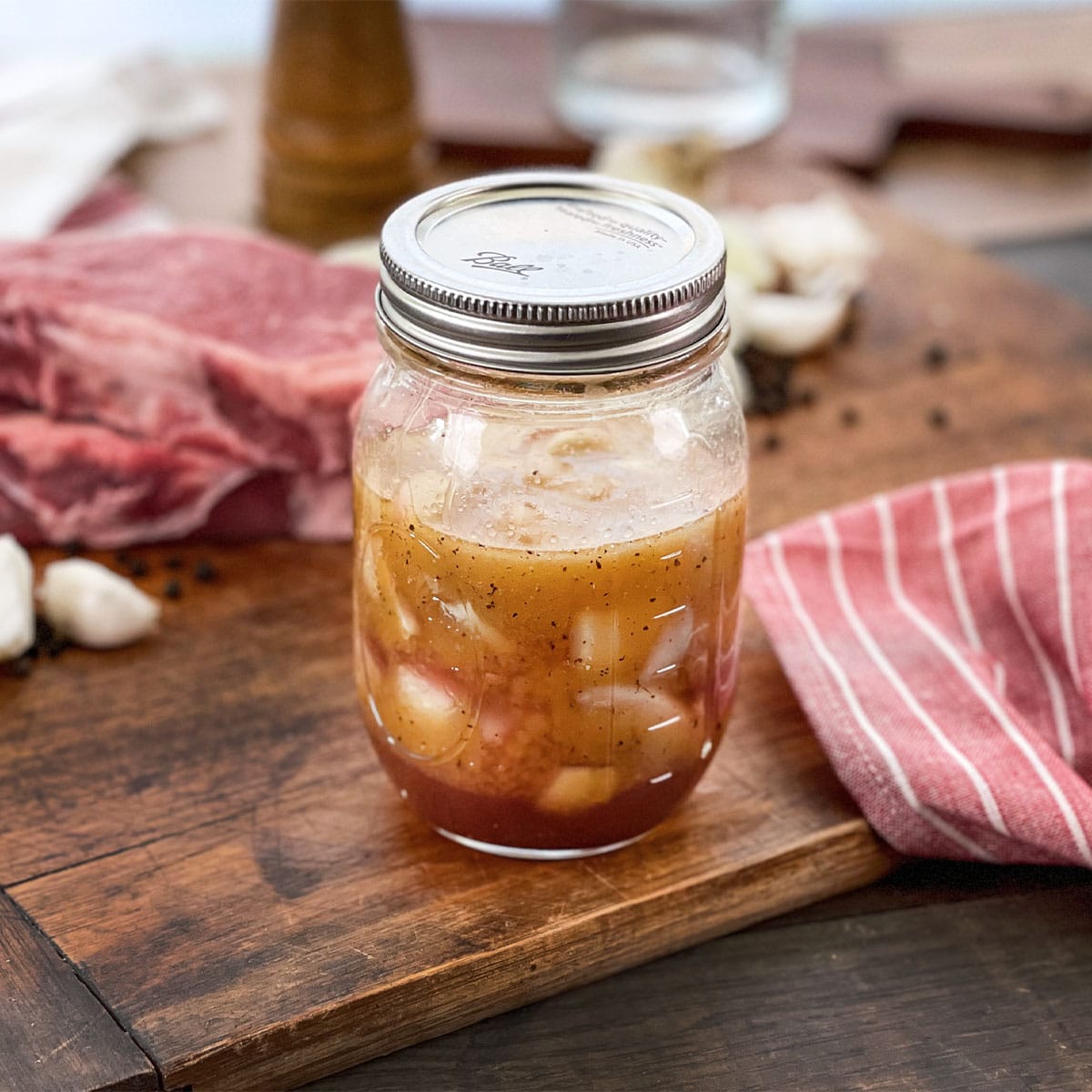 Mason jar with steak marinade with onion and garlic, raw steak in background.