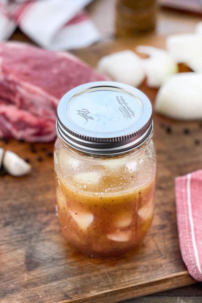 Mason jar with steak marinade with onion and garlic, raw steak in background.