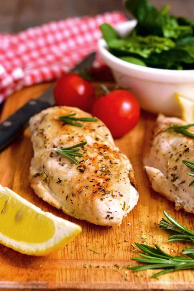 Baked chicken breast on a wooden board with lemon, herbs, and tomatoes.