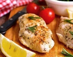 Baked chicken breast on a wooden board with lemon, herbs, and tomatoes.