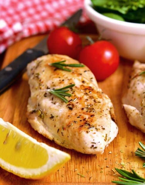Baked chicken breast on a wooden board with lemon, herbs, and tomatoes.