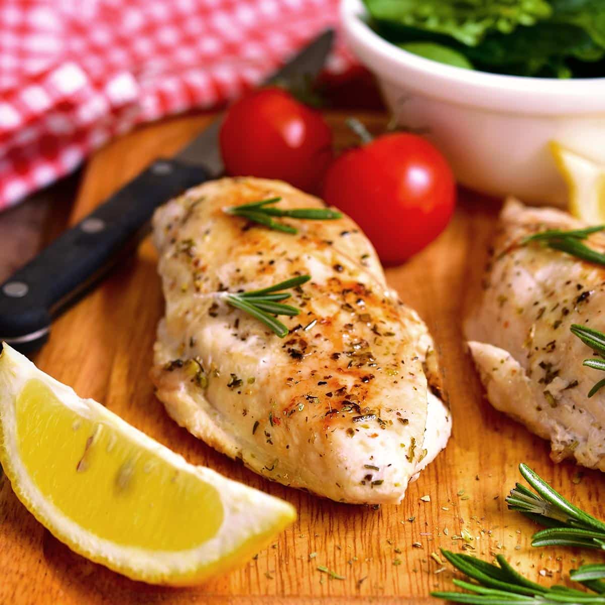 Baked chicken breast on a wooden board with lemon, herbs, and tomatoes.