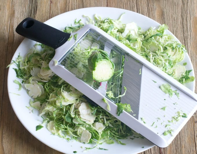 A large plate with shredded Brussels sprouts on it. A mandolin is lying on top of the shredded sprouts with a whole Brussels sprout on top ready to be shredded.