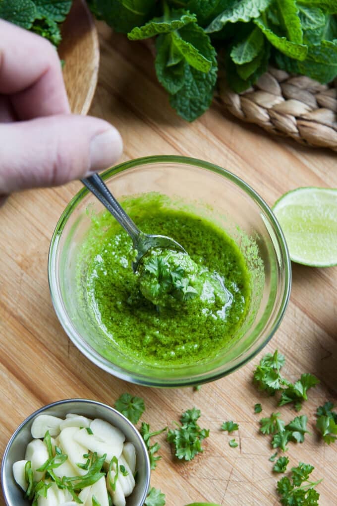 Glass bowl of homemade mint sauce with spoon, fresh ingredients in background.