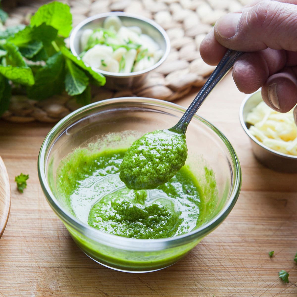 Glass bowl of homemade mint sauce with spoon, fresh ingredients in background.
