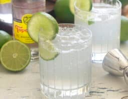 Ranch water cocktial in a low ball glass surrounded by limes and bottles of topo chico