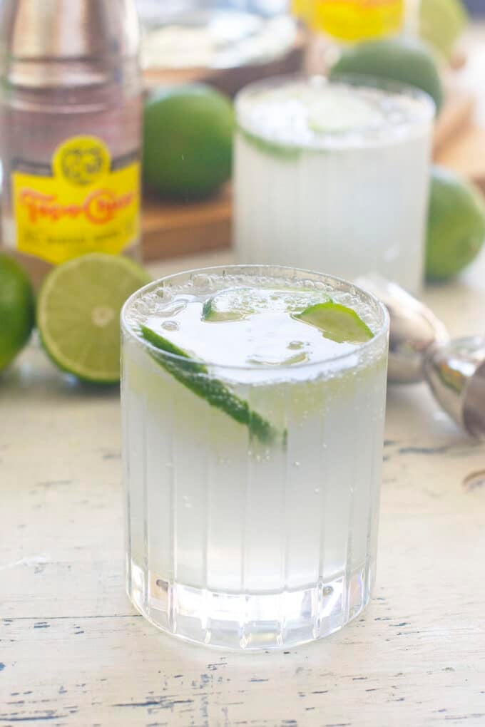 A lowball glass with a clear liquid, ranch water, a lime slice floating in it with bottles of topo chico and limes in the background.