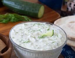 Glass bowl with tzatziki sauce, cucumbers in background.