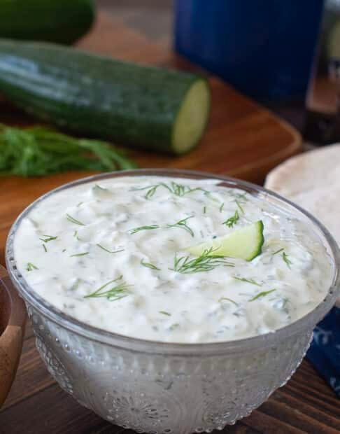 Glass bowl with tzatziki sauce, cucumbers in background.