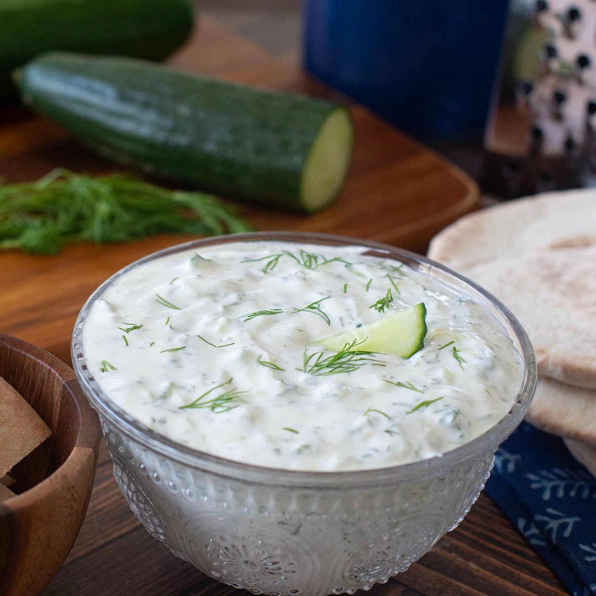 Glass bowl with tzatziki sauce, cucumbers in background.
