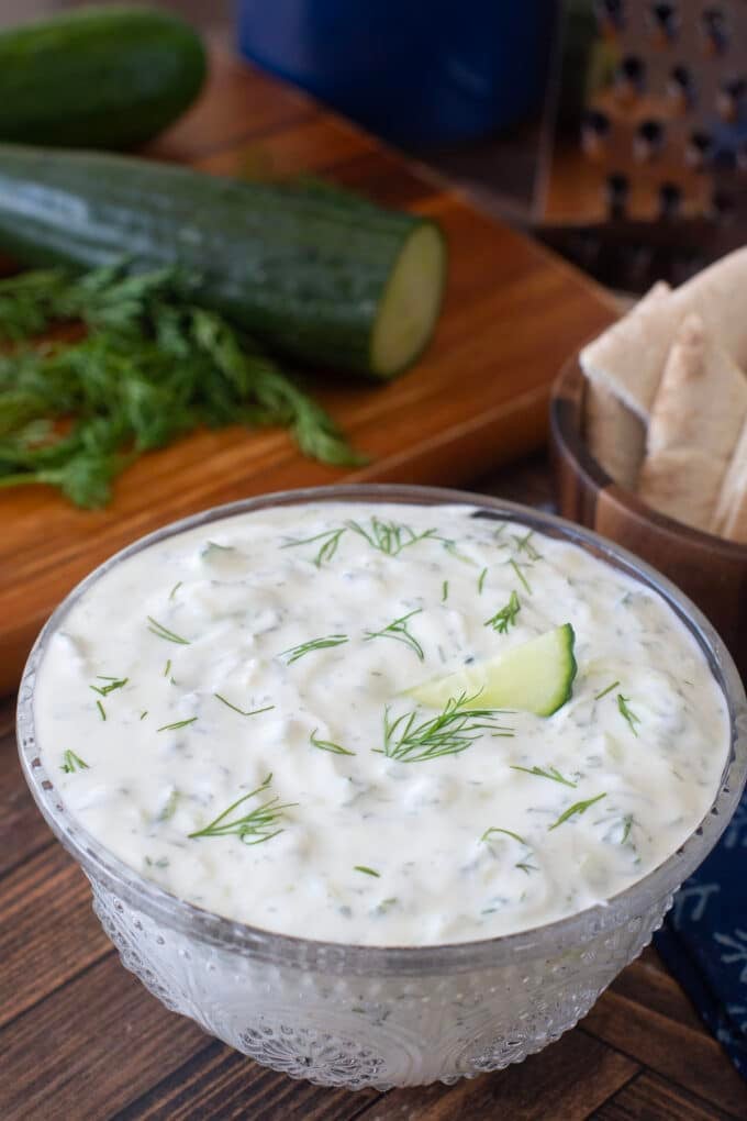 Glass bowl with tzatziki sauce, cucumbers in background.