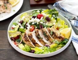 Greek salad with grilled and sliced chicken on top, lemons in background.