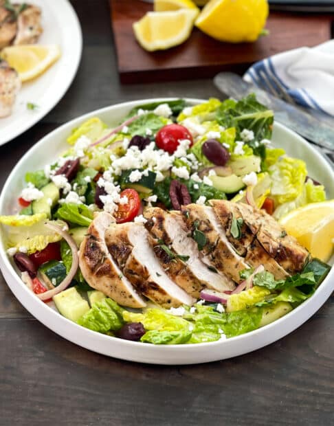 Greek salad with grilled and sliced chicken on top, lemons in background.