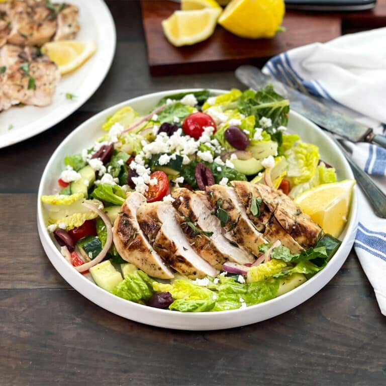 Greek salad with grilled and sliced chicken on top, lemons in background.