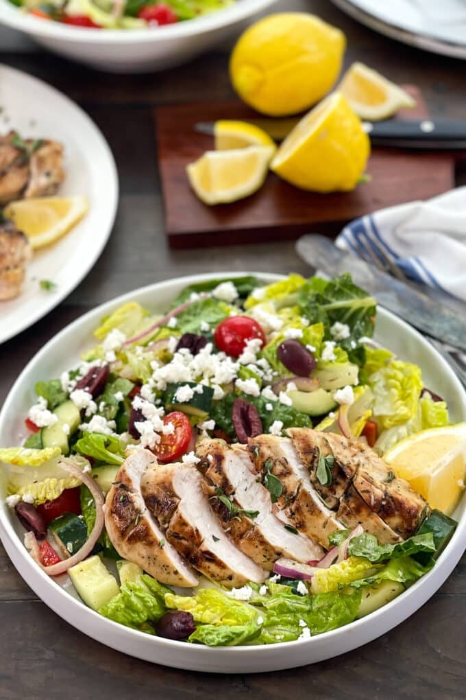 Greek salad with grilled and sliced chicken on top, lemons in background.