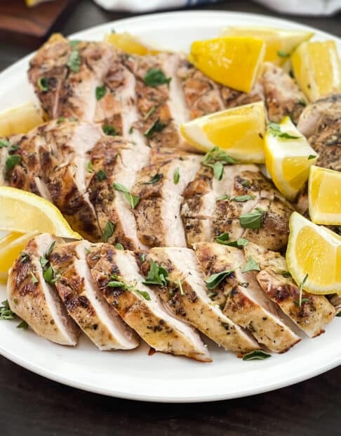 White platter of grilled and sliced chicken breasts with lemon wedges.