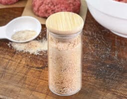 Jar of burger seasoning with raw burger patties in background.