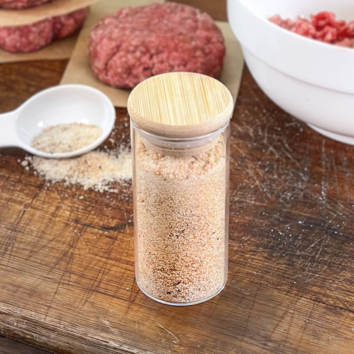 Jar of burger seasoning with raw burger patties in background.