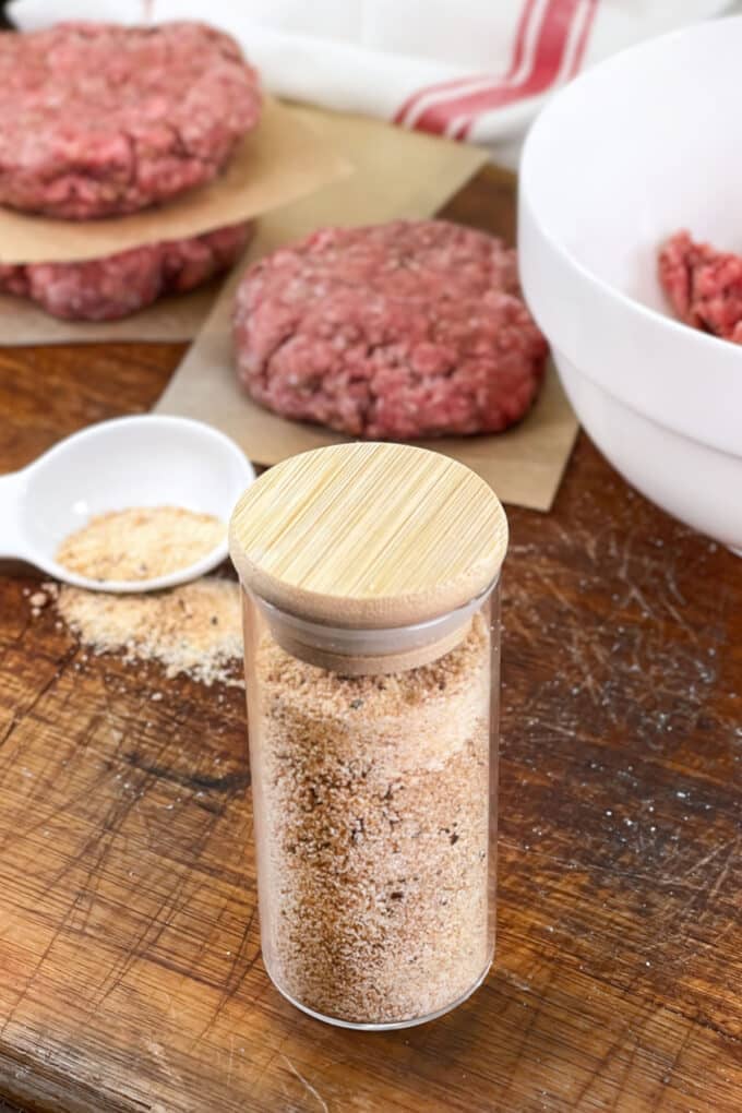 Jar of burger seasoning with raw burger patties in background.