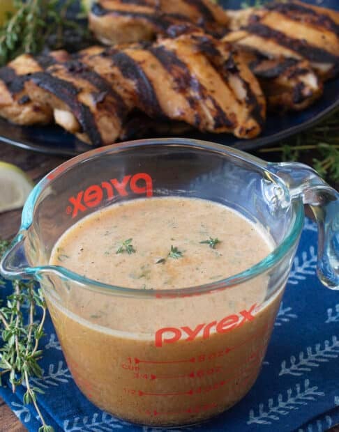 Glass measuring cup of chicken marinade with grilled chicken in background.