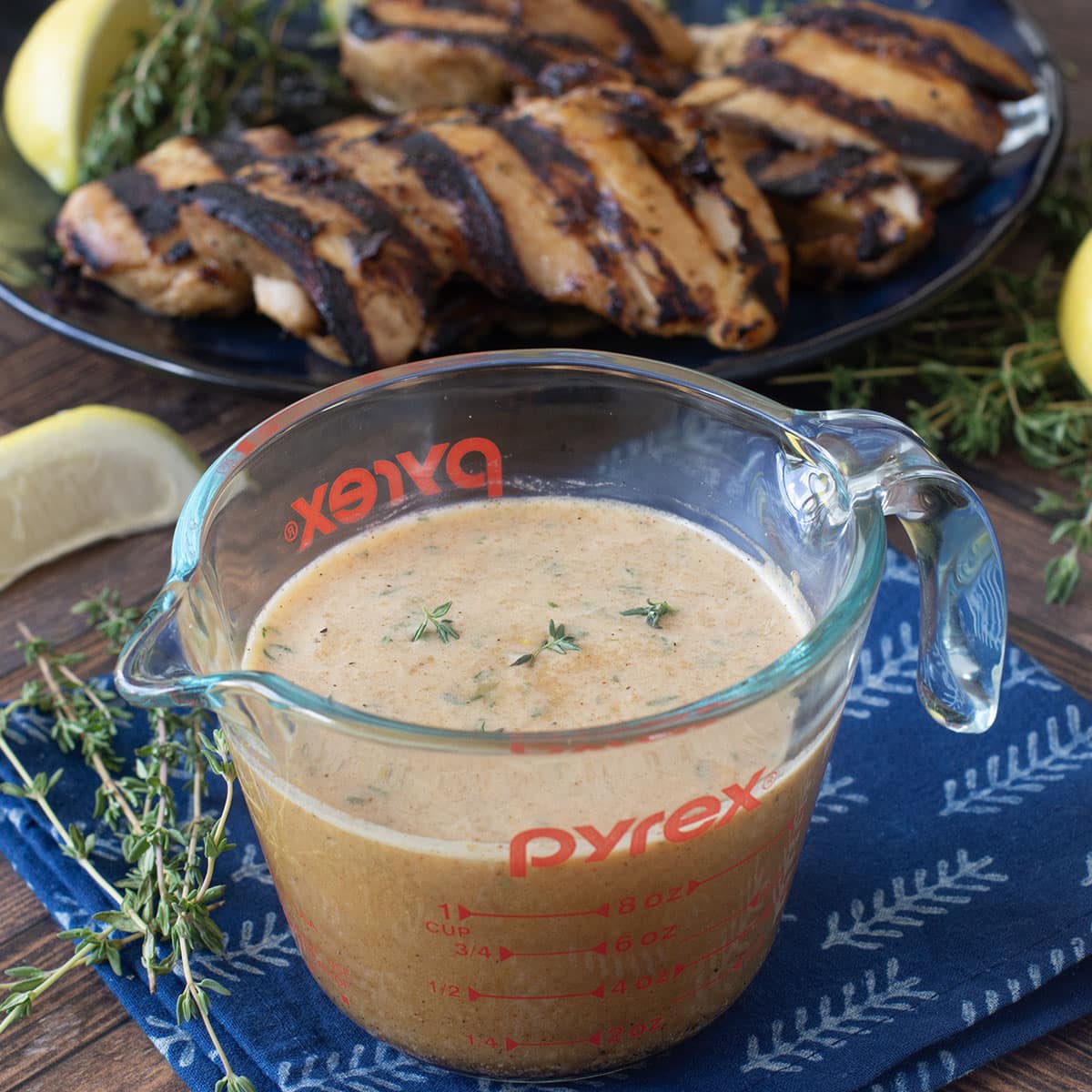 Glass measuring cup of chicken marinade with grilled chicken in background.
