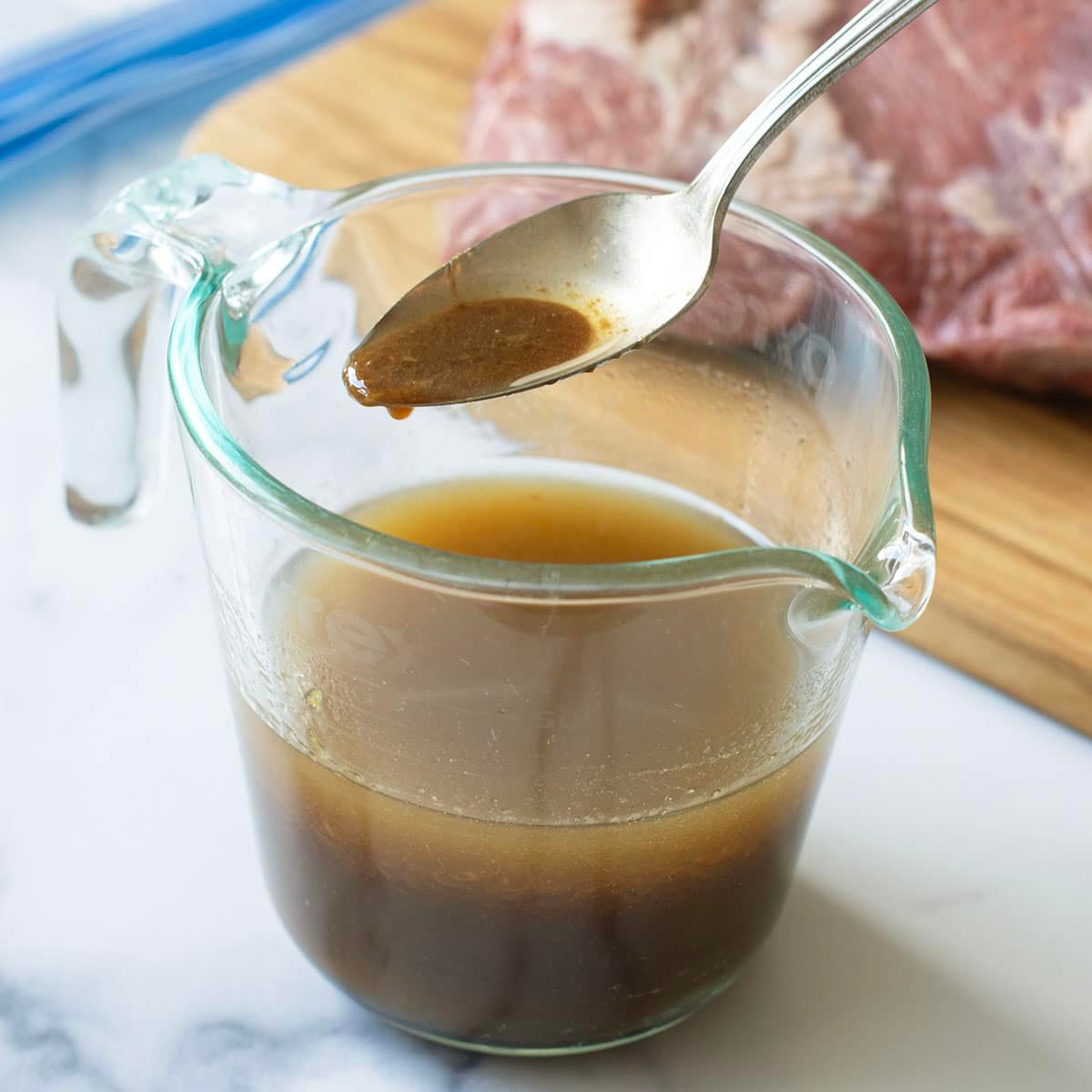 Glass measuring cup with brown marinade, spoonful above it, raw tri-tip in background.