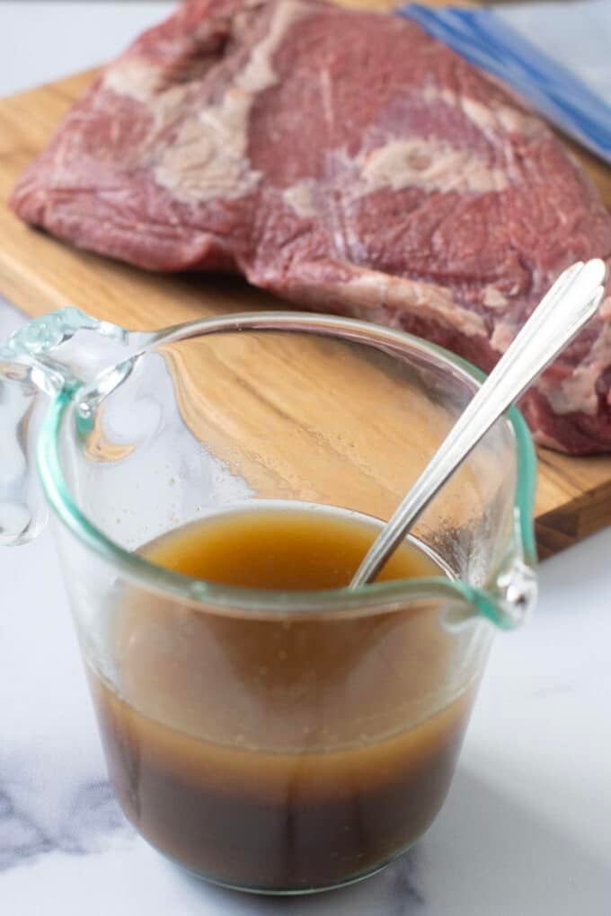 Glass measuring cup with brown marinade and spoon in it, raw tri-tip in background.